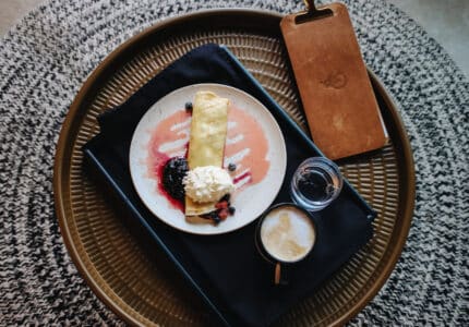 A plate with a crepe topped with berry sauce and whipped cream sits on a black cloth. Next to it are a small glass and a cup of coffee with milk foam on top. A wooden board rests nearby, all placed on a round woven surface.