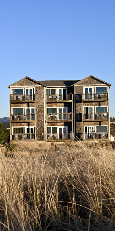 A multi-story building with a wooden facade is situated behind tall grass under a clear blue sky. Surrounding the building are green bushes and trees.