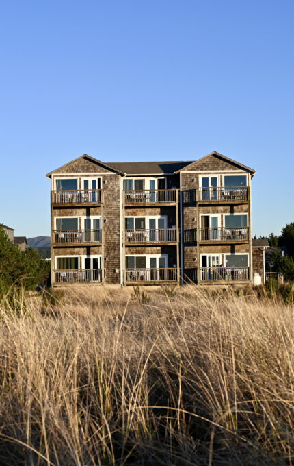 A multi-story building with a wooden facade is situated behind tall grass under a clear blue sky. Surrounding the building are green bushes and trees.
