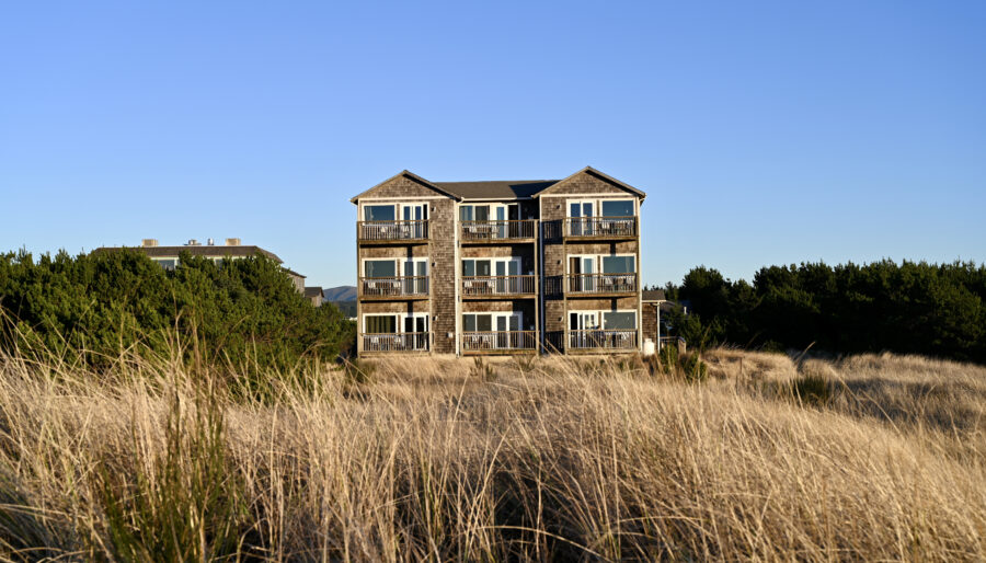 A multi-story building with a wooden facade is situated behind tall grass under a clear blue sky. Surrounding the building are green bushes and trees.