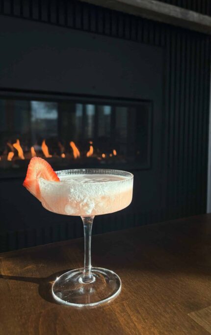 A cocktail in a glass with a sugar rim and a strawberry slice garnish sits on a wooden table. In the background, a lit fireplace adds a warm ambiance.