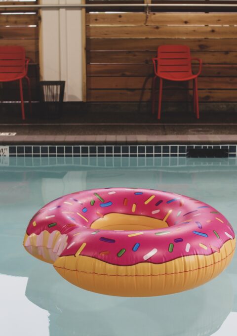 A swimming pool with a partially eaten donut-shaped inflatable float featuring pink frosting and colorful sprinkles. The pool has tiled edges and red chairs and a lifebuoy are visible on the deck.