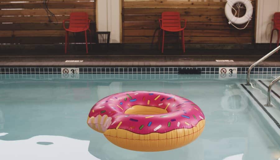 A swimming pool with a partially eaten donut-shaped inflatable float featuring pink frosting and colorful sprinkles. The pool has tiled edges and red chairs and a lifebuoy are visible on the deck.