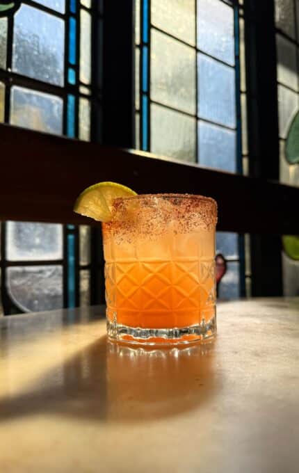 A glass with an orange cocktail, chili rim, and a lime wedge garnish sits on a table. Sunlight streams through stained glass windows in the background, casting shadows and colorful reflections.