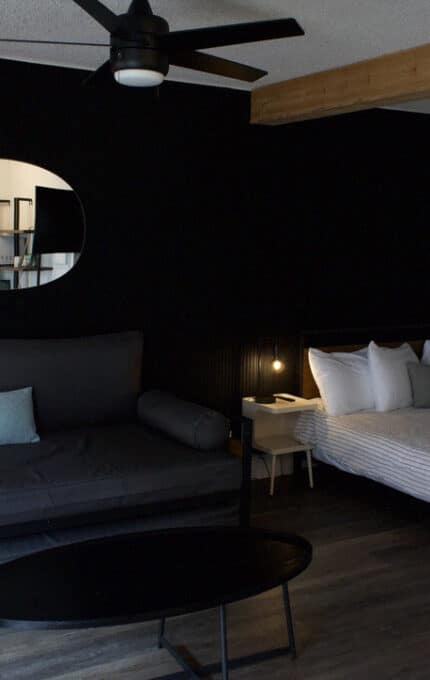 A modern bedroom with a black accent wall, a black sofa, a round mirror, a bed with white linens, two nightstands, a ceiling fan, and a round coffee table on a dark wood floor.