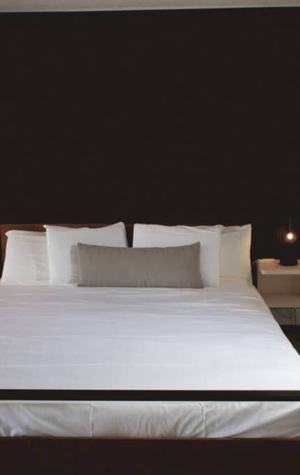 A neatly made bed with white linens and a gray pillow is centered in a modern room with a dark accent wall, two small white nightstands, hanging lamps, and a window with beige curtains on the right.