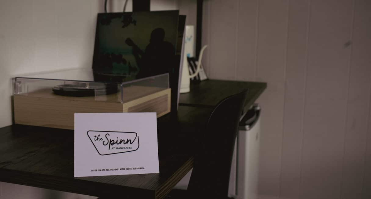 A white card labeled “the Spinn by Avamere” sits on a dark wooden desk next to a turntable, with a laptop and office supplies in the background. The wall is white with vertical paneling.