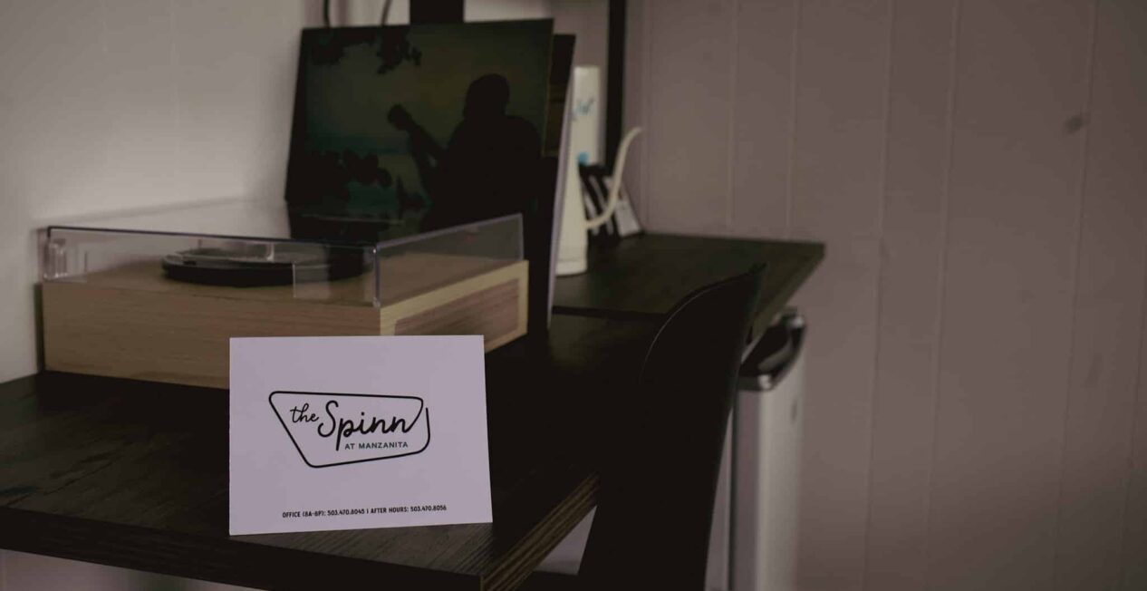 A white card labeled “the Spinn by Avamere” sits on a dark wooden desk next to a turntable, with a laptop and office supplies in the background. The wall is white with vertical paneling.