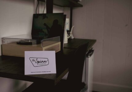 A white card labeled “the Spinn by Avamere” sits on a dark wooden desk next to a turntable, with a laptop and office supplies in the background. The wall is white with vertical paneling.