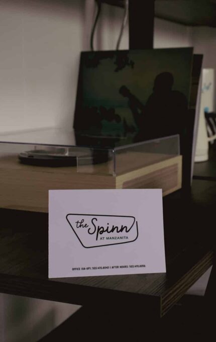A card with the text "the Spinn by Maximin" is displayed on a dark wooden desk. Behind it are a turntable, a vinyl record, a small fridge, and shelves against a white paneled wall.