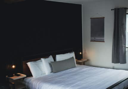 A neatly made bed with white linens and several pillows is centered in a room with dark and light walls. Two small nightstands with lamps flank the bed. A gray curtain covers a window, and a framed wall art hangs nearby.