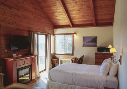 A cozy hotel room with wood-paneled walls and ceiling, a large bed, a small table with two chairs by the window, a fireplace, a flat-screen TV, and natural light coming through sliding glass doors.
