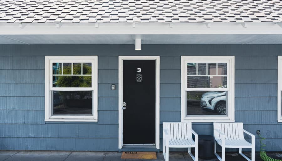 A black door with the number 3 and a “hello” doormat is centered between two white-framed windows on a blue building. Two white chairs sit to the right of the door. A car is partially visible through the right window.