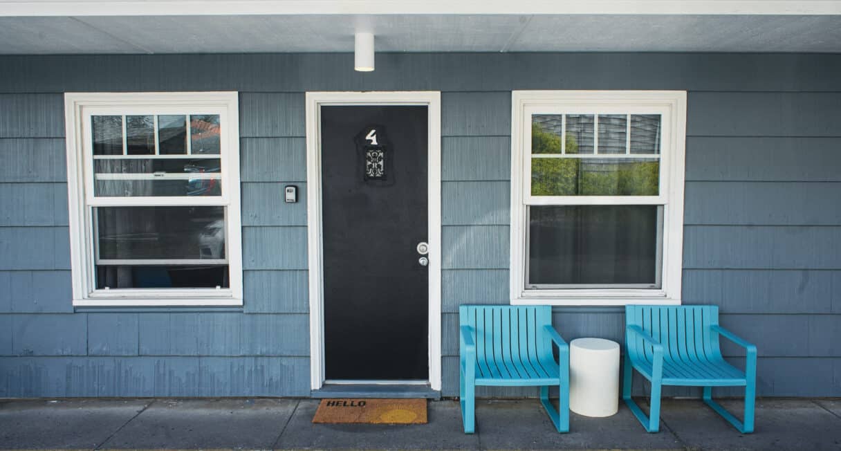 A blue-paneled building exterior with a black door labeled "4," flanked by two windows. Two turquoise chairs and a small white table sit on the right, and an orange "HELLO" doormat is in front of the door.
