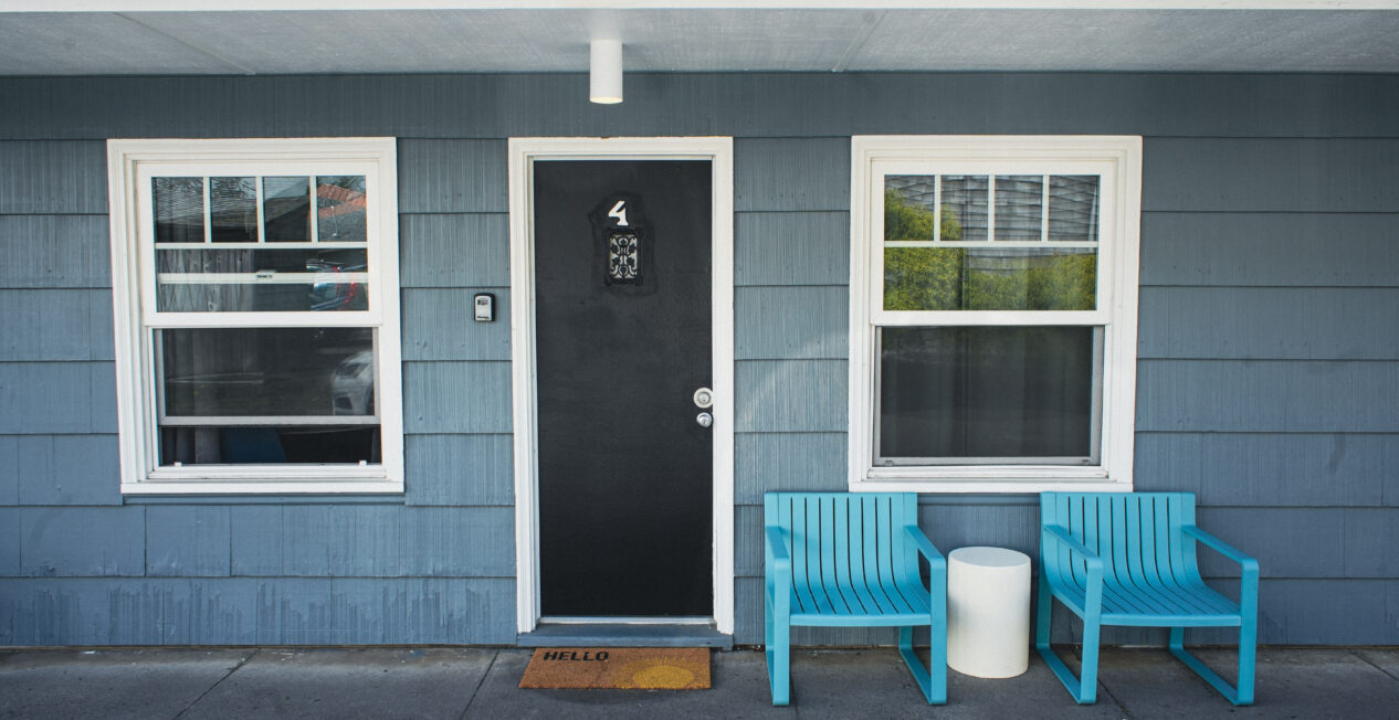 A blue-paneled building exterior with a black door labeled "4," flanked by two windows. Two turquoise chairs and a small white table sit on the right, and an orange "HELLO" doormat is in front of the door.