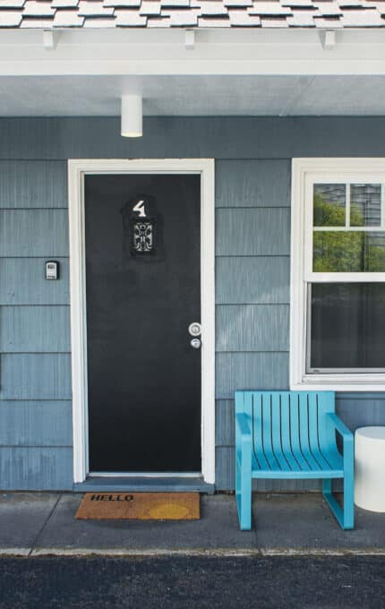 A blue-paneled building exterior with a black door labeled "4," flanked by two windows. Two turquoise chairs and a small white table sit on the right, and an orange "HELLO" doormat is in front of the door.
