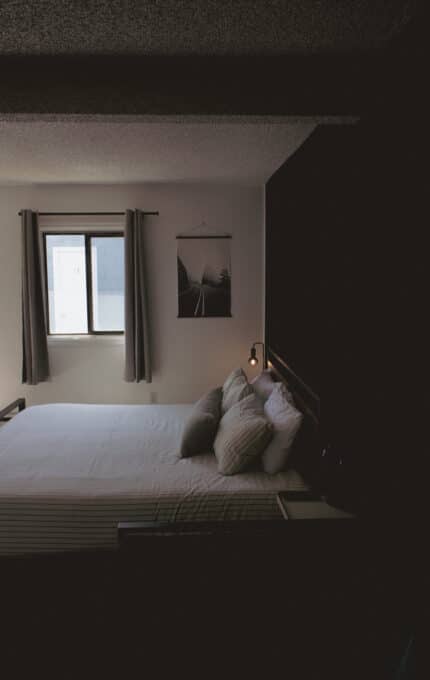 A neatly made bed with striped sheets and several pillows is placed against a dark wall. A small lamp is mounted to the wall above the bed, and a window with gray curtains lets in natural light.