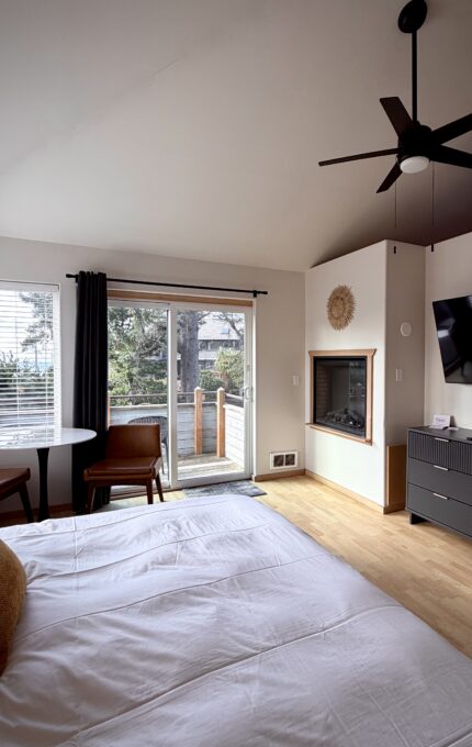 A modern bedroom with a neatly made bed, two chairs and a small round table by a window, a dresser with a TV above it, a fireplace in the corner, and a sliding door opening to a balcony with trees outside.