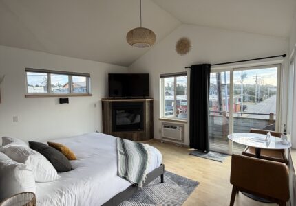A modern bedroom with a neatly made bed, a small round bedside table, a fireplace, a TV, a desk with a chair by large windows, and light wood flooring. Curtains are partly open, showing a view outside.