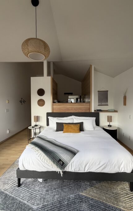 A modern bedroom with a neatly made bed, white bedding, and a mustard cushion. Two lamps sit on bedside tables. The space features wood accents, wall decor, a hanging wicker light, and an open layout with natural light.