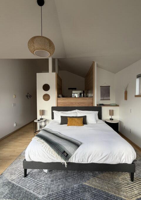 A modern bedroom with a neatly made bed, white bedding, and a mustard cushion. Two lamps sit on bedside tables. The space features wood accents, wall decor, a hanging wicker light, and an open layout with natural light.
