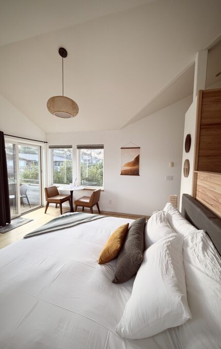 A bedroom with a neatly made bed, white linens, and accent pillows. There is a small dining table with two chairs by large windows. Natural light fills the room, and a wall hanging and pendant light are visible.