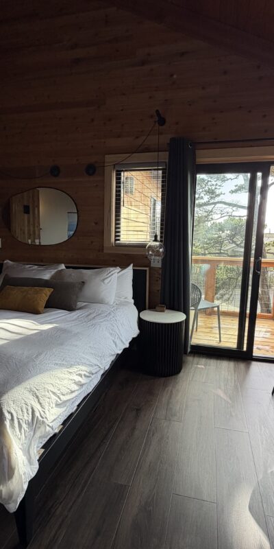 A cozy bedroom with wood-paneled walls and ceiling, a large bed with white bedding, a round mirror, a small table with two chairs, and a glass door leading to a balcony with outdoor seating and trees visible outside.