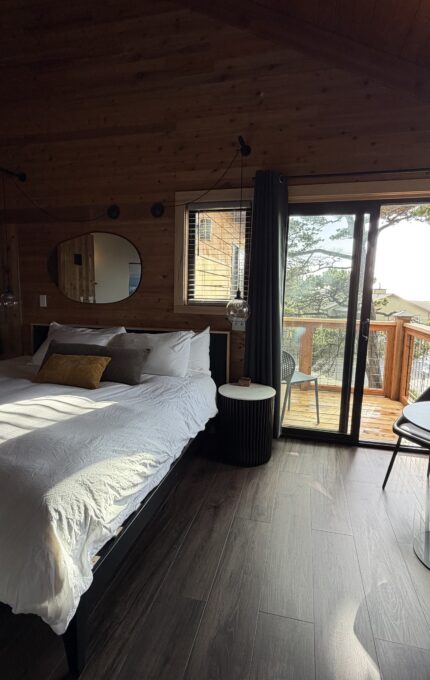 A cozy bedroom with wood-paneled walls and ceiling, a large bed with white bedding, a round mirror, a small table with two chairs, and a glass door leading to a balcony with outdoor seating and trees visible outside.