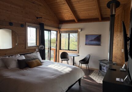 A cozy bedroom with wood-paneled walls and ceiling, a large bed with white bedding, a small table with two chairs near the window, a wall mirror, a wood stove, and natural light coming through windows.