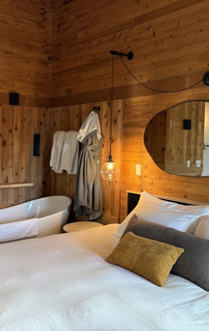 A cozy wooden bedroom features a neatly made bed with white sheets and accent pillows, a freestanding bathtub with a towel, bathrobes hanging on the wall, a round mirror, and a round light fixture.