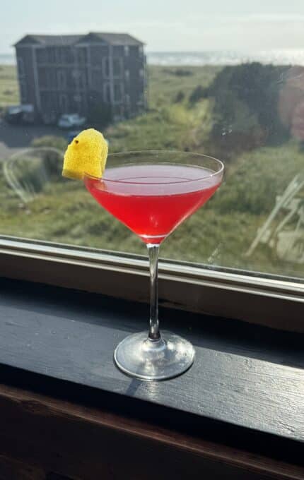 A bright pink cocktail in a martini glass, garnished with a lemon twist, sits on a windowsill with a grassy landscape, a distant building, and blue sky visible outside.
