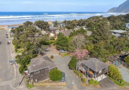 Aerial view of a coastal neighborhood with houses, trees, parked cars, and roads near a sandy beach and the ocean; a forested hill rises in the background under a partly cloudy sky.