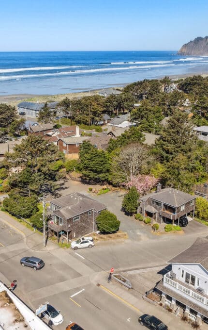 Aerial view of a coastal town with houses, trees, and roads near a sandy beach and the ocean, with forested hills in the background under a clear blue sky.