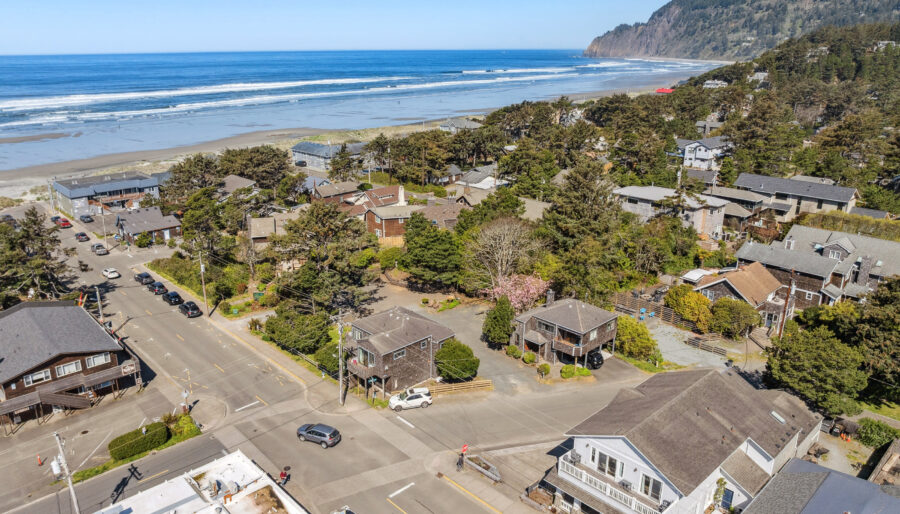 Aerial view of a coastal town with houses, trees, and roads near a sandy beach and the ocean, with forested hills in the background under a clear blue sky.