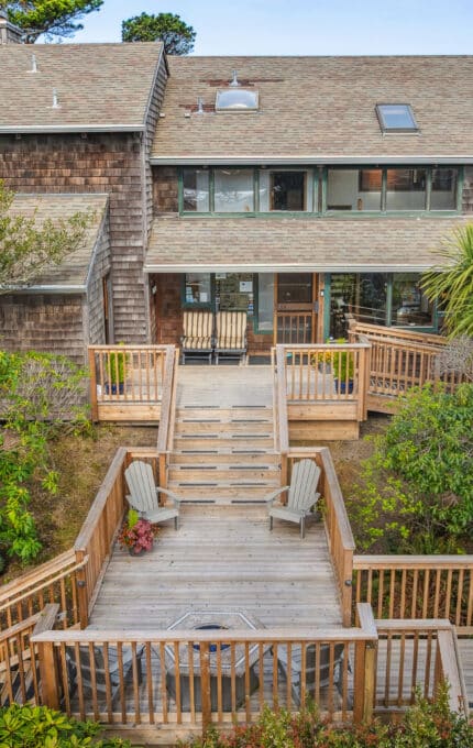A two-story wooden house with large windows and a shingle exterior features multiple decks connected by stairs, surrounded by greenery and outdoor seating in the foreground.