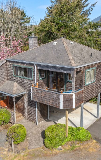 A two-story house with weathered wood siding stands elevated on stilts. There is a covered carport beneath, a small porch, trees and bushes around the property, and neighboring houses in the background.