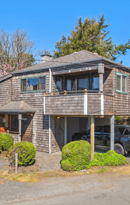 A two-story house with wooden shingle siding and a raised deck, surrounded by green bushes. An SUV is parked underneath the deck. The sky is clear and blue, with trees in the background.