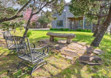 A wooden bench and two metal chairs sit on a stone patio under trees in a grassy yard, with a shingled house and blooming trees visible in the background.