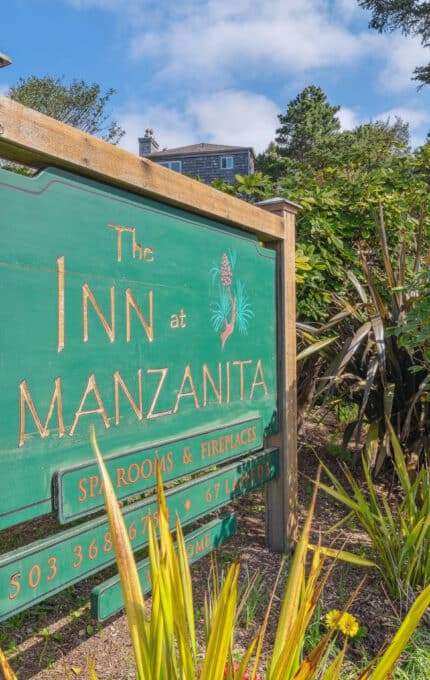 A large wooden sign for The Inn at Manzanita stands near wooden stairs and lush greenery. The sign displays contact information and advertises spa rooms and fireplaces. Trees and a building appear in the background.