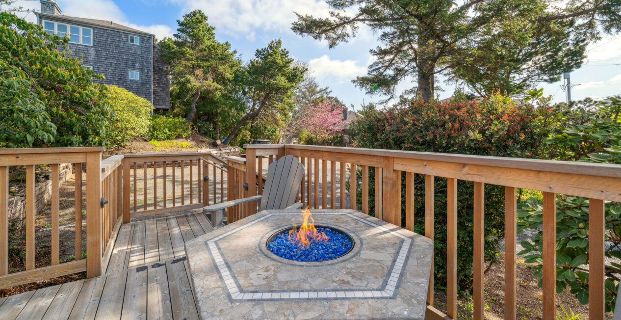 A wooden deck with a stone fire pit table surrounded by Adirondack chairs, enclosed by a wooden railing, and surrounded by trees and shrubs under a partly cloudy sky.