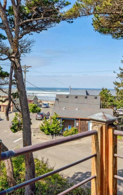 View from a wooden balcony overlooking a street lined with trees and houses, with the ocean and waves visible in the background under a clear blue sky.