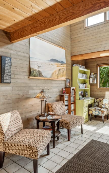 A sunlit sitting area with two patterned chairs, a small table, a lamp, wall art, bookshelves, and large windows overlooking greenery, all set against wooden walls and a tiled floor.