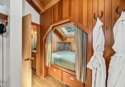 A cozy bedroom with wood panel walls features a bed beneath a skylight, curtains for privacy, storage drawers under the bed, and two white robes hanging on hooks on the wall. An open door leads to a bathroom.
