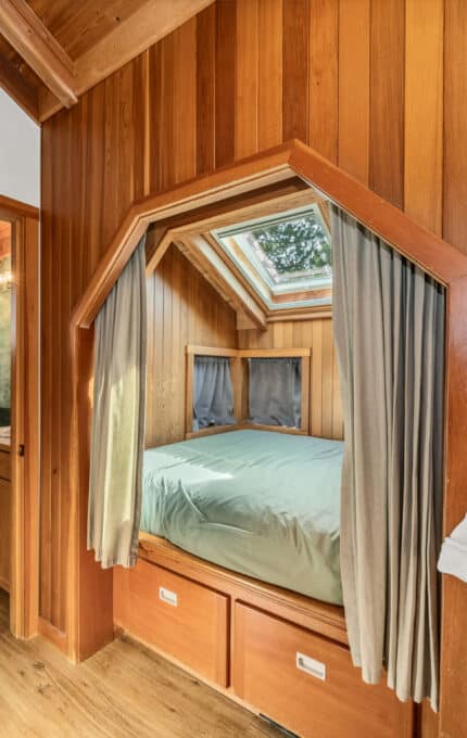 A cozy bedroom with wood panel walls features a bed beneath a skylight, curtains for privacy, storage drawers under the bed, and two white robes hanging on hooks on the wall. An open door leads to a bathroom.