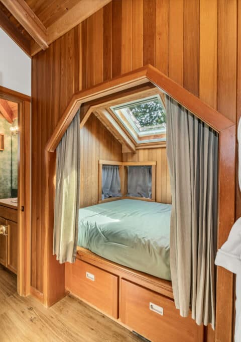 A cozy bedroom with wood panel walls features a bed beneath a skylight, curtains for privacy, storage drawers under the bed, and two white robes hanging on hooks on the wall. An open door leads to a bathroom.