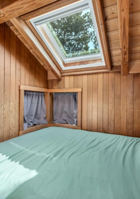 A cozy bedroom with wood-paneled walls and ceiling, featuring a skylight above the bed and two small curtained windows. The bed has a green comforter and a decorative pillow. Natural light enters the room.