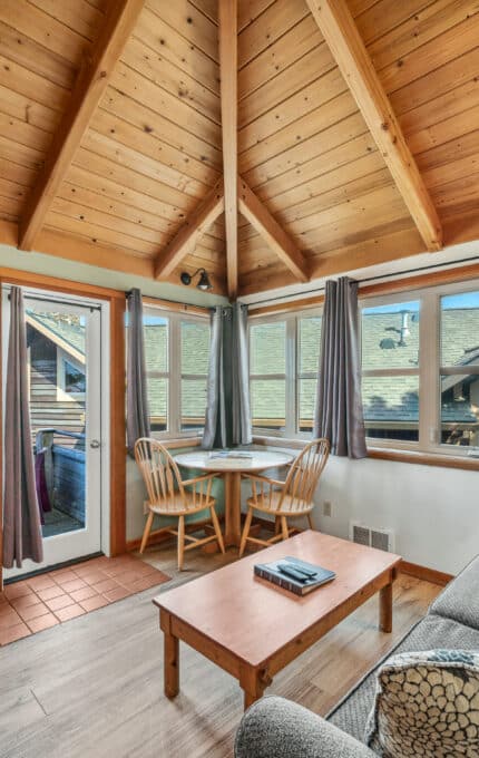 A cozy living room with a wooden vaulted ceiling, gray sofa, small wooden coffee table, dining nook with two chairs, wall-mounted TV, and a fireplace. Large windows and a glass door let in natural light.