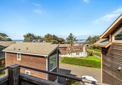 View from a balcony overlooking wooden houses and a quiet street, with trees and the ocean visible in the background under a clear blue sky.
