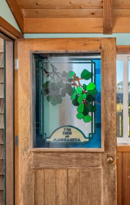 A wooden door with a stained glass window showing green leaves and the words "The Inn Alexandra." The door is in a wood-paneled entryway with a window and partial outdoor view of trees.