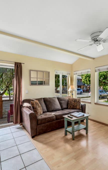 A small living room with a brown sofa, light wood coffee table, and side table. There is a dining area with a round table and two chairs by sliding glass doors. Large windows allow natural light to fill the space.